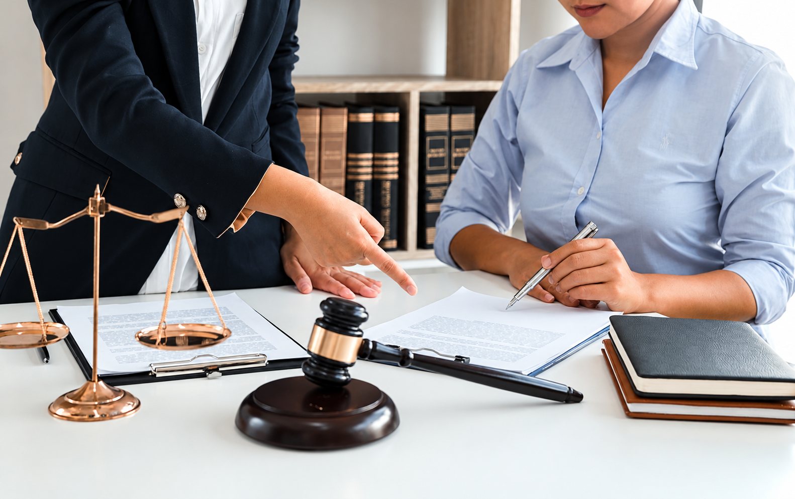 Deux femmes en tenue professionnelle examinent un document sur un bureau où sont posés une balance de la justice, un marteau de juge et des carnets.