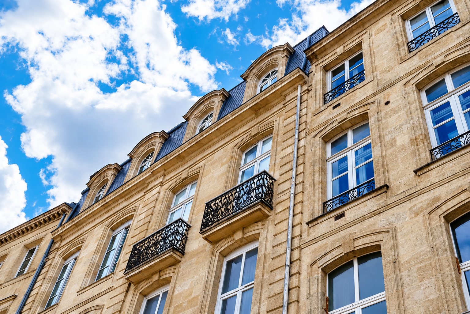Vue en contre-plongée d'une façade en pierre de taille avec balcons en fer forgé, lucarnes sur toit d'ardoise et ciel bleu nuageux.