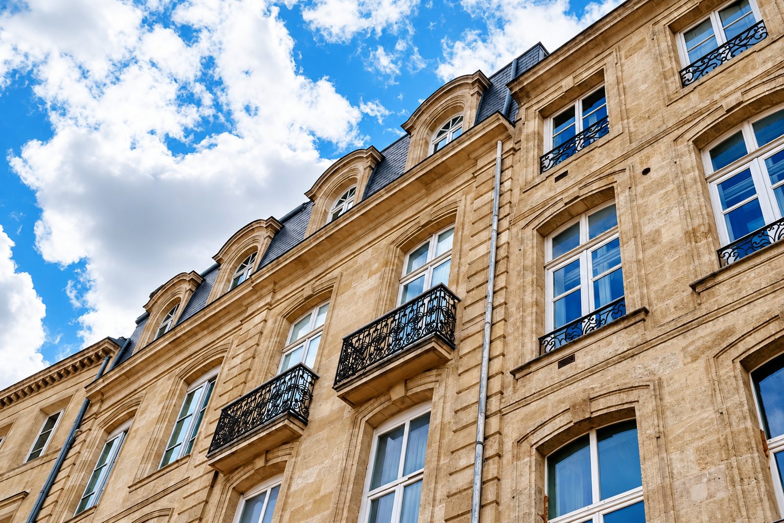 Façade d'un immeuble ancien en pierre blonde avec balcons en fer forgé, lucarnes et toit d'ardoise sous un ciel bleu nuageux.