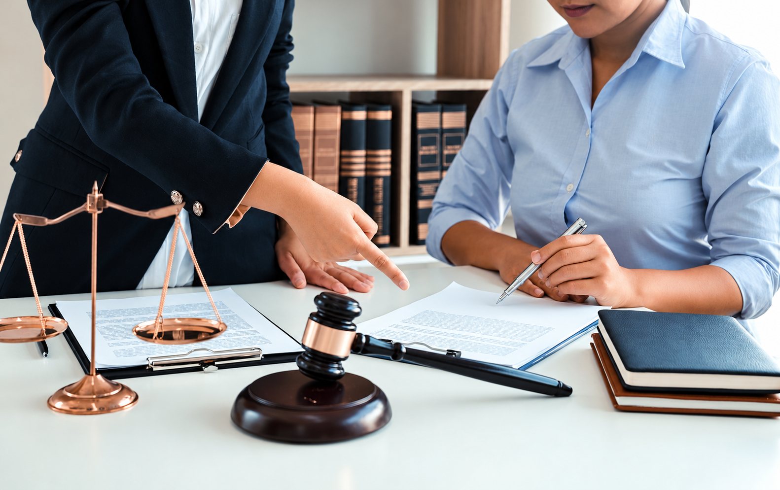 Une avocate en veste sombre désigne un document pendant qu'une cliente en chemise bleue signe, sur un bureau avec balance de la justice et marteau de juge.
